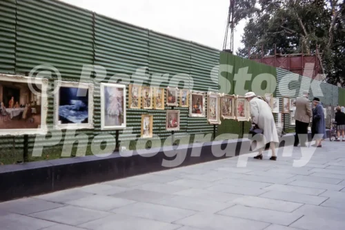 A 1950s colour photo of an outdoor art exhibition along a corrugated green fence on Piccadilly in London, with pedestrians in mid-century attire pausing to view the framed paintings.