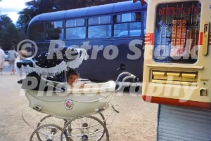 A 1950s colour photo of a young child in a vintage cream-coloured pram parked next to a Nestlé’s chocolate vending machine, with a blue bus in the background.