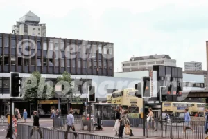 A 1970s street scene at Ironmonger Row in Coventry featuring double-decker buses, traffic lights, and pedestrians near modern office buildings.