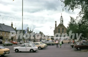 A 1970s view of the High Street in Moreton-in-Marsh featuring several period cars, a large coach, and the historic Curfew Tower under a cloudy sky.