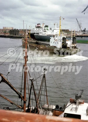 A 1970s view of activity at the Newcastle docks on the River Tyne, featuring several large industrial vessels and the stern of a smaller boat in the foreground.