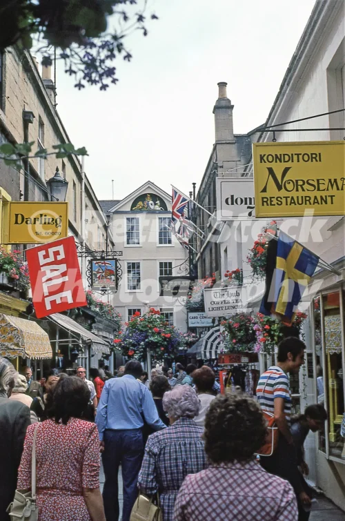 1980s photo of Northumberland Place in Bath with crowded shoppers, hanging flower baskets, sale signs and shop signs including Darling and Konditori Norseman Restaurant.
