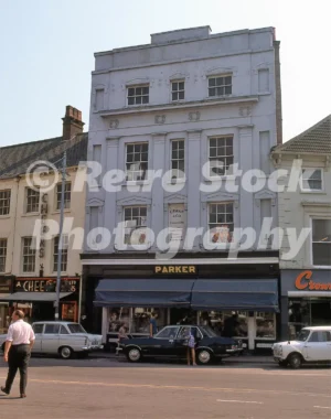 A 1970s photograph of the C. Parker & Co. stationery shop at 19 Market Place in Louth, Lincolnshire, featuring a blue sun awning and several classic cars including a white Mini and a dark saloon parked on the street.