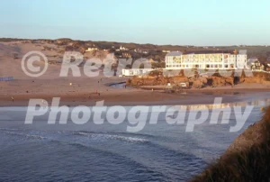 A 1970s photograph of Perranporth Beach in Cornwall at dusk, showing the large white Perran Sands Hotel situated on the cliffs overlooking the beach and sand dunes.