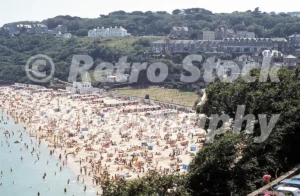 A 1970s photograph looking down over a crowded Porthminster Beach in St Ives, Cornwall, showing hundreds of holidaymakers, colourful windbreaks, and the wooded hillside with Victorian villas in the background.