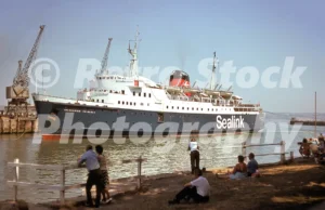 A 1975 photograph of the Sealink ferry 'Caledonian Princess' at Weymouth Harbour, featuring its blue and white hull, red funnel, and onlookers sitting on a grassy bank behind a white fence.