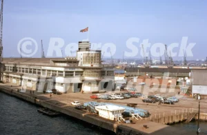 A 1972 photograph of the Art Deco Ocean Terminal in Southampton, featuring the curved passenger gallery with a Union Jack flag flying above, a car park with period vehicles, and industrial cranes in the background.