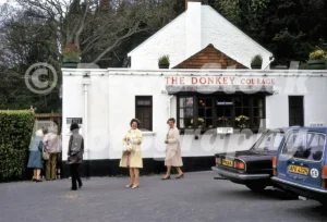 A 1971 photograph of The Donkey public house in Farnham, Surrey, featuring two women in coats standing in the car park next to a black Triumph and a blue Volvo estate.