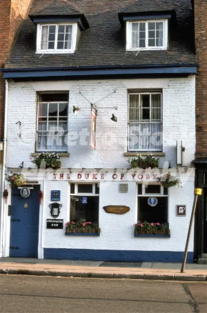 A 1975 photograph of The Duke of York public house on Barton Street in Tewkesbury, featuring the white-painted facade, blue doors and trim, and floral window boxes.