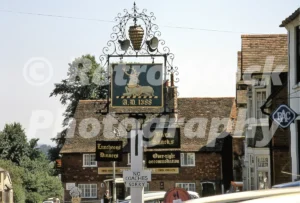 A 1970 photograph of the ornate hanging sign for The Prince Albert pub in Bletchingley, Surrey, featuring a white hart and A.D. 1388, with the timber-framed pub building and a "No Coaches Sorry" sign in the background.