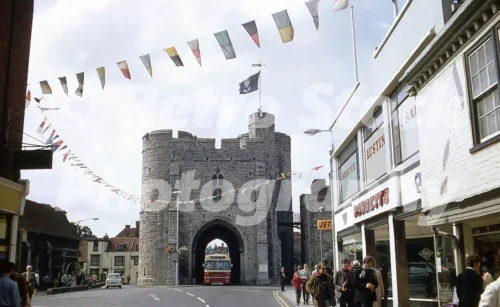 A 1974 photograph showing an East Kent A.E.C. Reliance bus passing through the narrow stone arch of the Westgate in Canterbury, with Barretts Austin car dealership visible on the right.
