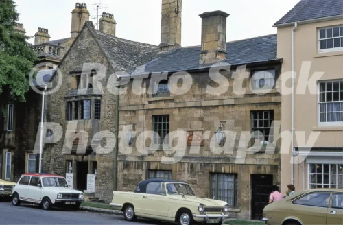 A 1978 photograph of the Woolstaplers Hall Museum in Chipping Campden, featuring the historic Cotswold stone building with a prominent gable, a white Mini Clubman, and a cream Triumph Herald convertible parked in front.
