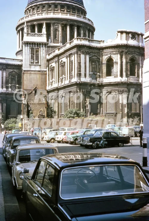A 1960s colour photo of a line of parked classic cars, on a street beside the soot-stained stone walls and dome of St Paul's Cathedral in London in 1964.