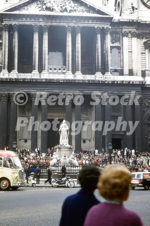 A 1960s colour photo of a crowd gathered in front of the soot-stained stone facade and Queen Anne statue of St Paul's Cathedral in London in 1961.