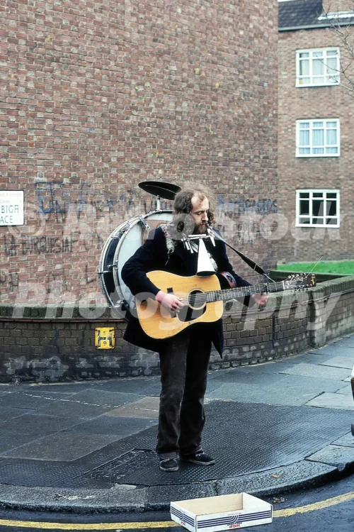1980s photo of a street busker in Denbigh Terrace, Notting Hill, London, playing guitar with a one-man-band drum kit on a street corner.