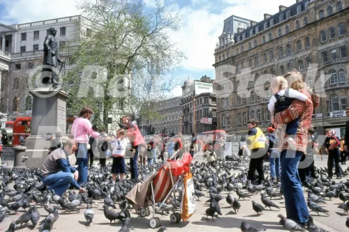 1970s photo of families and children feeding pigeons in Trafalgar Square with a pushchair, red buses and the Superama Shopping sign in the background