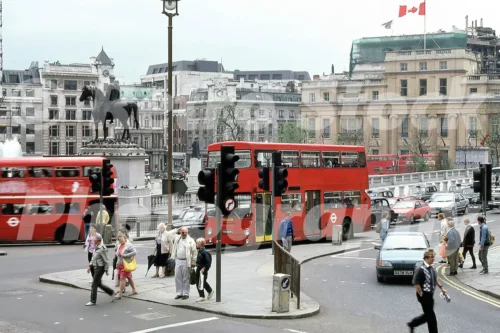 1980s photo of Trafalgar Square in London, with pedestrians crossing at a busy junction, red London buses including a Daimler Fleetline, and the equestrian statue overlooking the square.