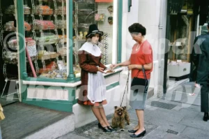 A 1965 photograph of the exterior of a traditional sweet shop at 2 High Street in Conwy, featuring a young woman in Welsh national costume offering samples to a customer with a small dog.