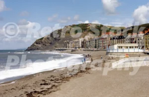 A 1977 photograph of the pebble beach and seafront at Aberystwyth, looking north toward Constitution Hill with the Cliff Railway visible and waves breaking on the shore.