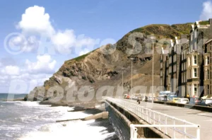 A 1977 photograph of the promenade in Aberystwyth, looking toward Constitution Hill with waves crashing against the sea wall and period cars parked alongside Victorian buildings.