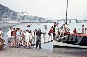 A 1960s photograph of a group of schoolboys in short trousers and jumpers boarding the Barmouth Ferry boat via a wooden plank on the sandy shore of the Mawddach Estuary.