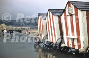 A 1950s photograph of traditional red and white striped wooden bathing machines on large wheels parked along the wet promenade in Llandudno, with a sign requesting the public not to feed seagulls.