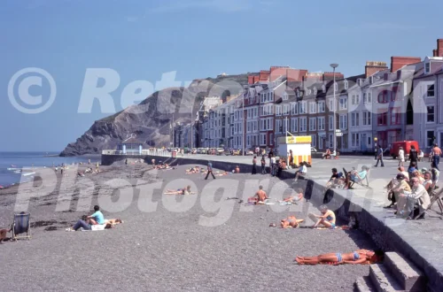 A 1977 photograph of sunbathers on the shingle beach at Aberystwyth, with the Victorian promenade, a yellow ice cream kiosk, and Constitution Hill in the background.