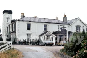 A 1977 photograph of the Chequers Hotel in Northop Hall, featuring the white Victorian manor house with a crenellated tower, a hanging sign, and a dark green Ford Granada parked out front.
