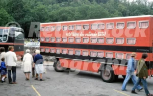 A 1979 view of a red Rotel Tours Mercedes-Benz bus with a large "Das Rollende Hotel" trailer parked on a narrow road in the Welsh countryside.