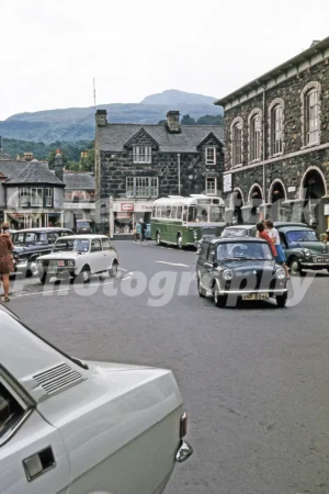 A 1970s view of Finsbury Square in Dolgellau featuring a green coach, several Mini cars, and traditional stone buildings under the backdrop of Cader Idris.