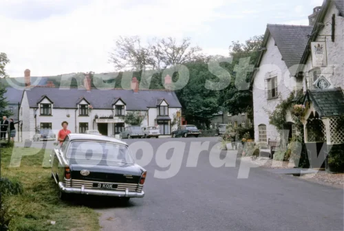 A 1960s view of the Hand Hotel and The West Arms in Llanarmon Dyffryn Ceiriog featuring a black Ford Zodiac with a GB sticker and several other period cars.