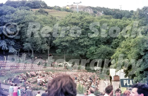 A 1960s view of the open-air Happy Valley Theatre in Llandudno, featuring rows of spectators in deckchairs watching a variety performance on a white stage.