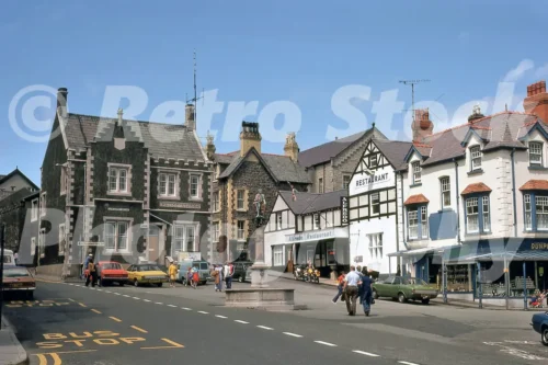 A 1970s street view of Lancaster Square in Conwy featuring the stone Police Station, Alfredo Restaurant, and the Prince Llywelyn monument with period cars.