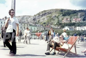 A 1960s view of Llandudno promenade featuring a man in a deckchair reading a newspaper, a Royal Navy Rescue helicopter in the background, and the Little Orme cliffs.