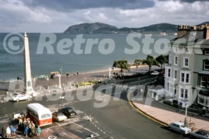 A 1970s elevated view of Llandudno Promenade featuring a red and white Crosville coach parked by the granite obelisk war memorial with the Little Orme in the background.