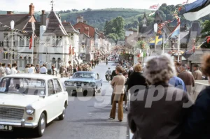 A 1969 street view of Castle Street in Llangollen crowded with people and international flags, featuring a rare blue grey Ford Mustang with registration GBP 315C.