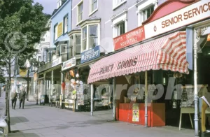 A 1967 photograph of the sunlit pavement on Mostyn Street in Llandudno, featuring several gift shops with striped awnings, a Hoover Service Depot, and pedestrians walking past displays of seaside souvenirs.