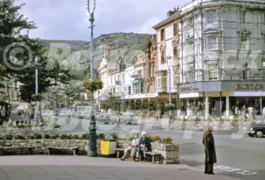 A 1967 street view of Mostyn Street in Llandudno featuring shoppers on a stone bench, a Burton tailoring store under scaffolding, and the limestone cliffs of the Great Orme.