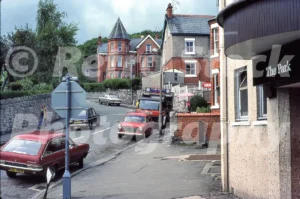 A 1979 photograph of Nant-y-Glyn Road in Colwyn Bay, featuring a red Vauxhall Viva, a red Austin 1100, and a large red truck, with Victorian brick villas in the background.