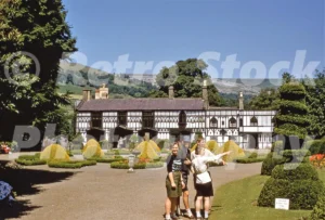 A 1950s photograph of Plas Newydd in Llangollen featuring the ornate black-and-white timbered house and formal gardens with topiary, while four young visitors stand in the foreground.