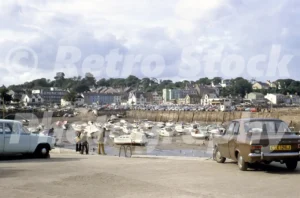 A 1970s photograph of Saundersfoot harbour at low tide, featuring dozens of small boats resting on the sand, a brown Ford Escort parked in the foreground, and the town rising in the background.