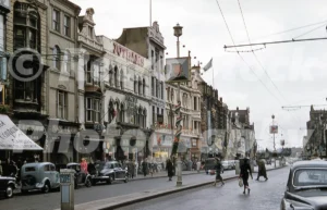 A 1950s photograph of St Mary Street in Cardiff, featuring the bustling city centre with double-decker buses, vintage black cars, and shoppers walking past historic storefronts like Jothams and Slade.
