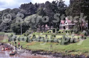 A 1970s photograph of the Ferry Hotel in Tal-y-cafn on the banks of the River Conwy, featuring the half-timbered building, tiered gardens with topiary, and several small boats moored at the stone slipway.