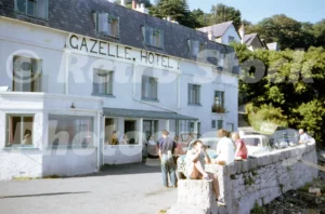 A 1960s photograph of the Gazelle Hotel in Glyn Garth, featuring the white waterfront building with "GAZELLE HOTEL" painted on the facade, people relaxing on a stone sea wall, and vintage cars parked along the narrow coastal road.