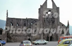 A 1970s photograph of the West Front of Tintern Abbey, featuring the intricate Gothic tracery of the great west window with several vintage cars parked in the foreground including a silver Renault 5 and a white Vauxhall Viva.