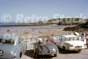 A 1960s photograph of Traeth Bychan beach at low tide, featuring several vintage cars parked on the shingle including a grey Morris Minor convertible and a white Ford Consul Classic, with small boats on the sand and the coastline in the distance.
