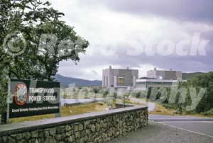 A 1960s photograph of the Trawsfynydd Nuclear Power Station in North Wales, featuring the prominent black and white entrance sign in the foreground and the two massive concrete reactor buildings in the distance under a cloudy sky.