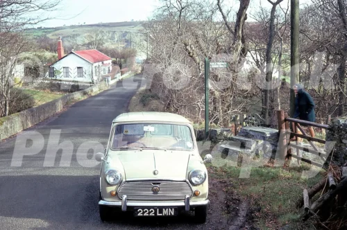 A 1976 colour photo of a cream-coloured Austin Mini (registration 222 LMN) parked on a narrow road leading to Nun Mill Bay, with a public footpath sign for "Dhoon Beach" and a white cottage with a red roof in the background.