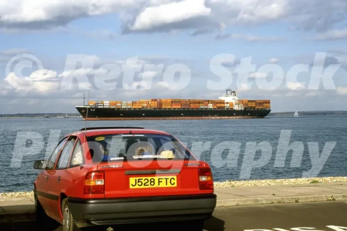 Red Vauxhall Cavalier Mk3 parked by the water in the 1990s with a large container ship passing in the background
