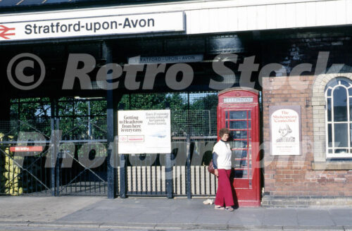 1970s view of Stratford-upon-Avon station in Warwickshire with British Rail signage, a red telephone box, entrance posters and a woman walking past the brick station frontage